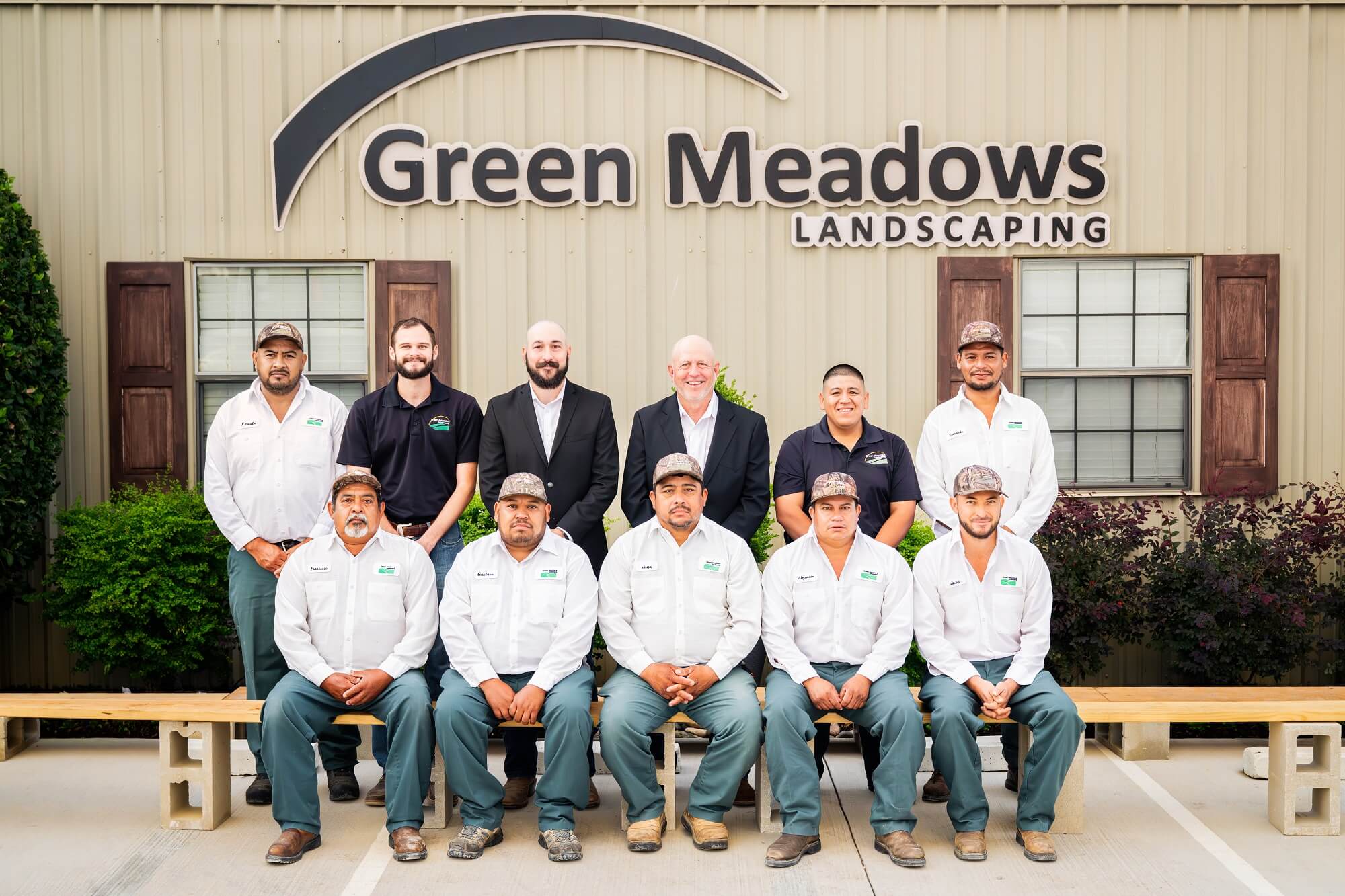 A group of twelve men, some in white work shirts and green pants, others in business attire, pose in front of a building with a sign that reads Green Meadows Landscaping.