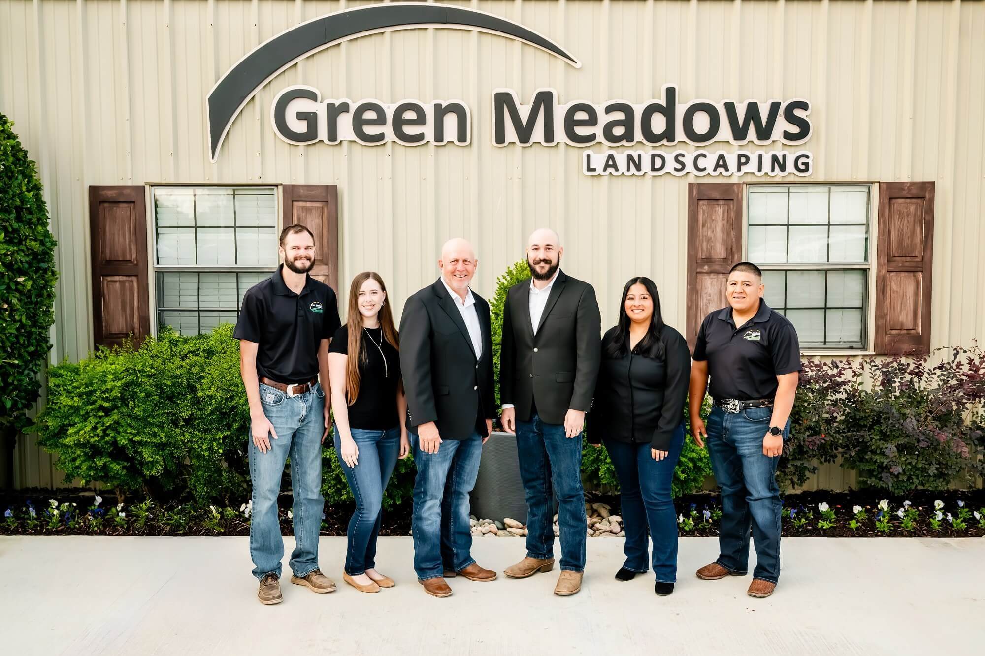 Six people stand smiling in front of a Green Meadows Landscaping building. Four wear black company shirts and jeans, while two men in the center wear suits. Green shrubs and flowers line the building.