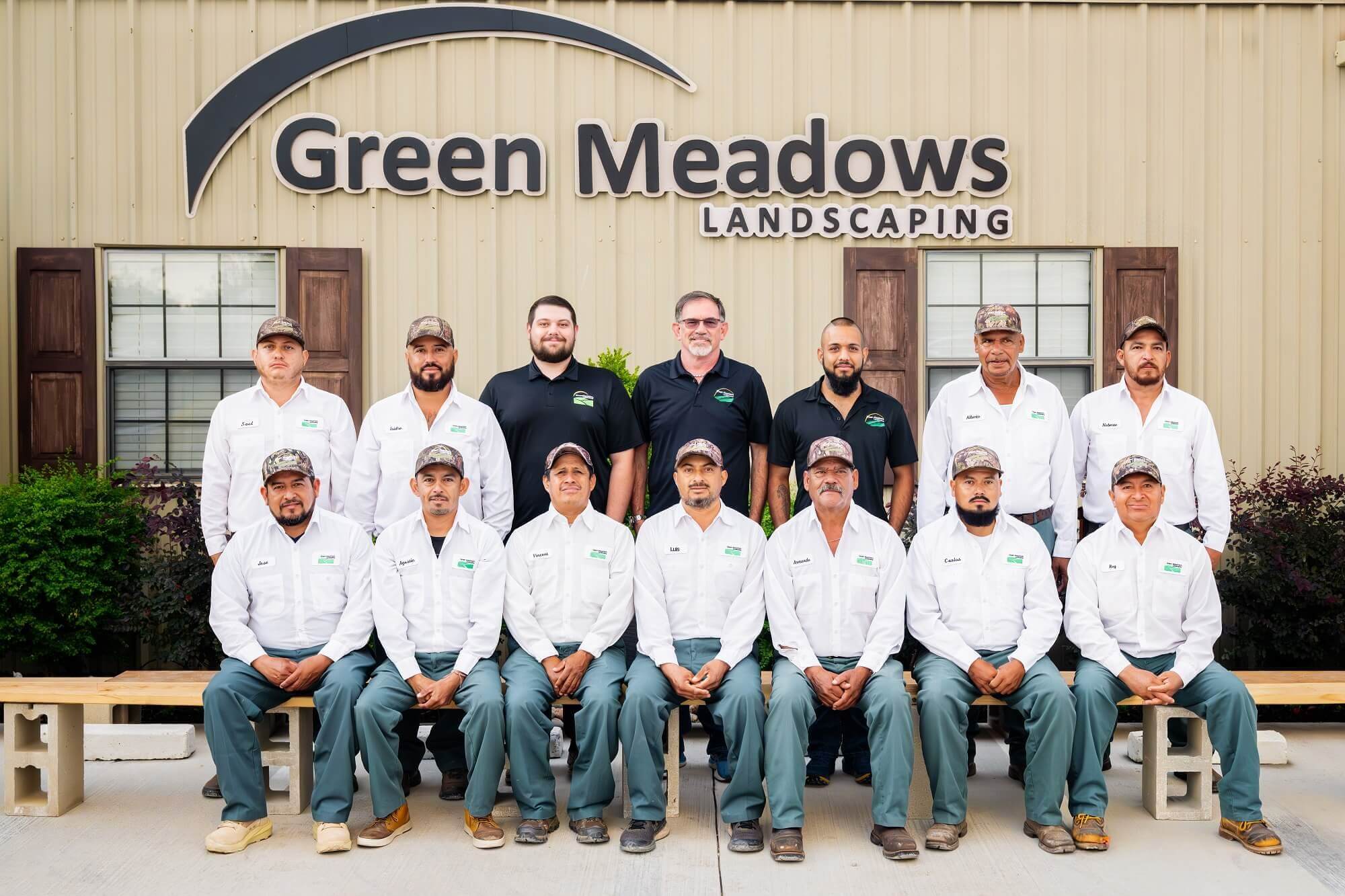 A group of fourteen men, some seated and some standing, pose in front of a building with a sign that reads Green Meadows Landscaping. Most wear white shirts and green pants; three wear black shirts.