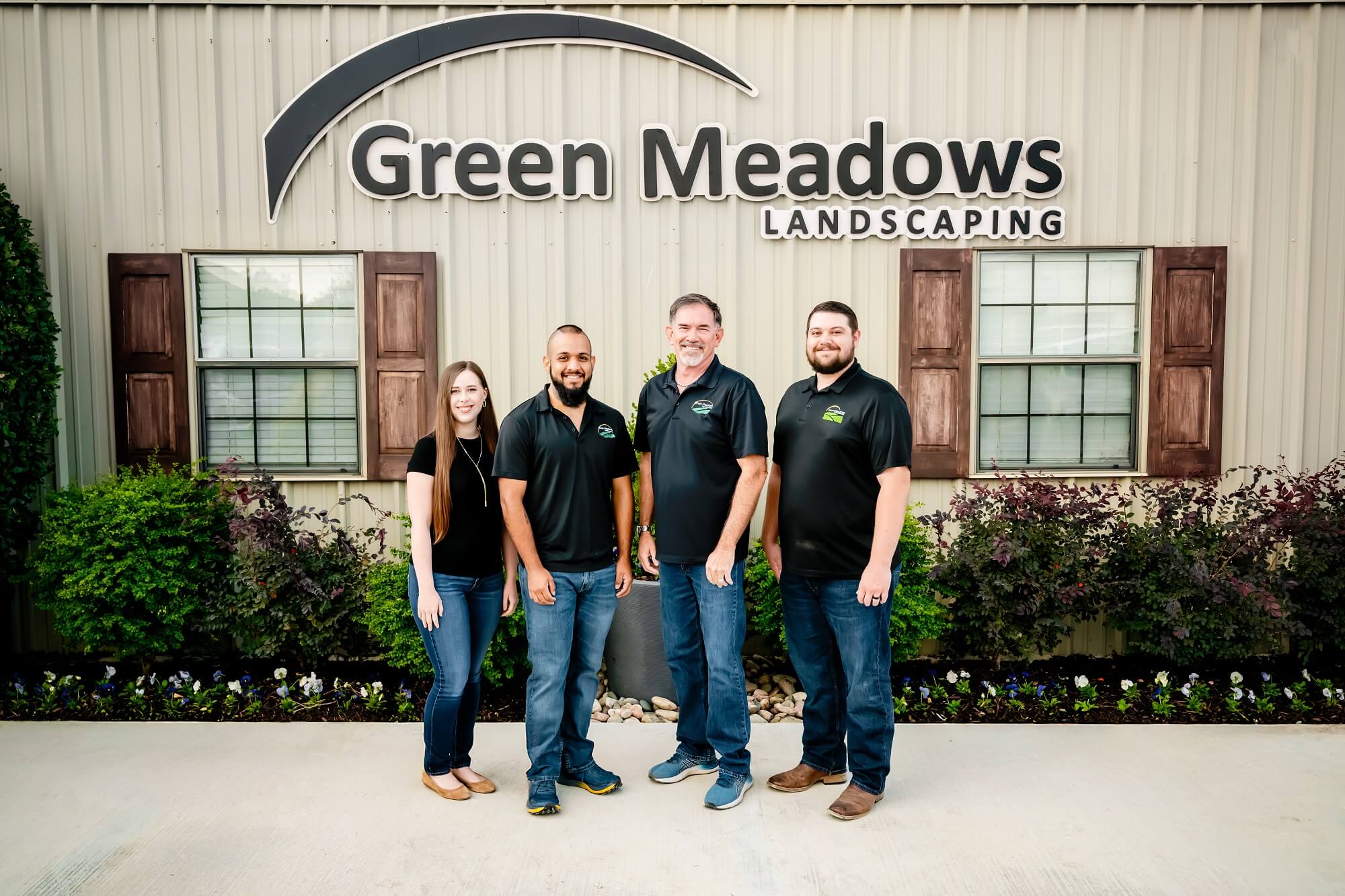Four people stand smiling in front of a building with a Green Meadows Landscaping sign, two windows with brown shutters, and landscaping plants along the wall. All wear black shirts and jeans or khakis.
