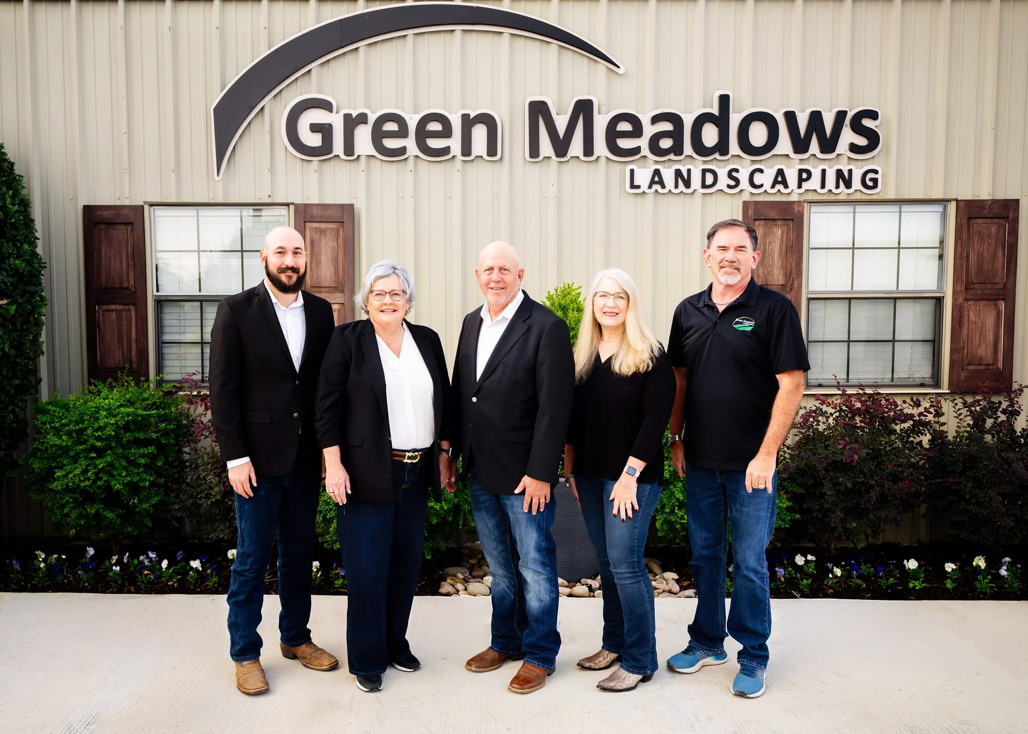 Five people stand smiling in front of a building with a sign reading Green Meadows Landscaping. They are dressed in business casual attire, with greenery and flowers visible behind them.
