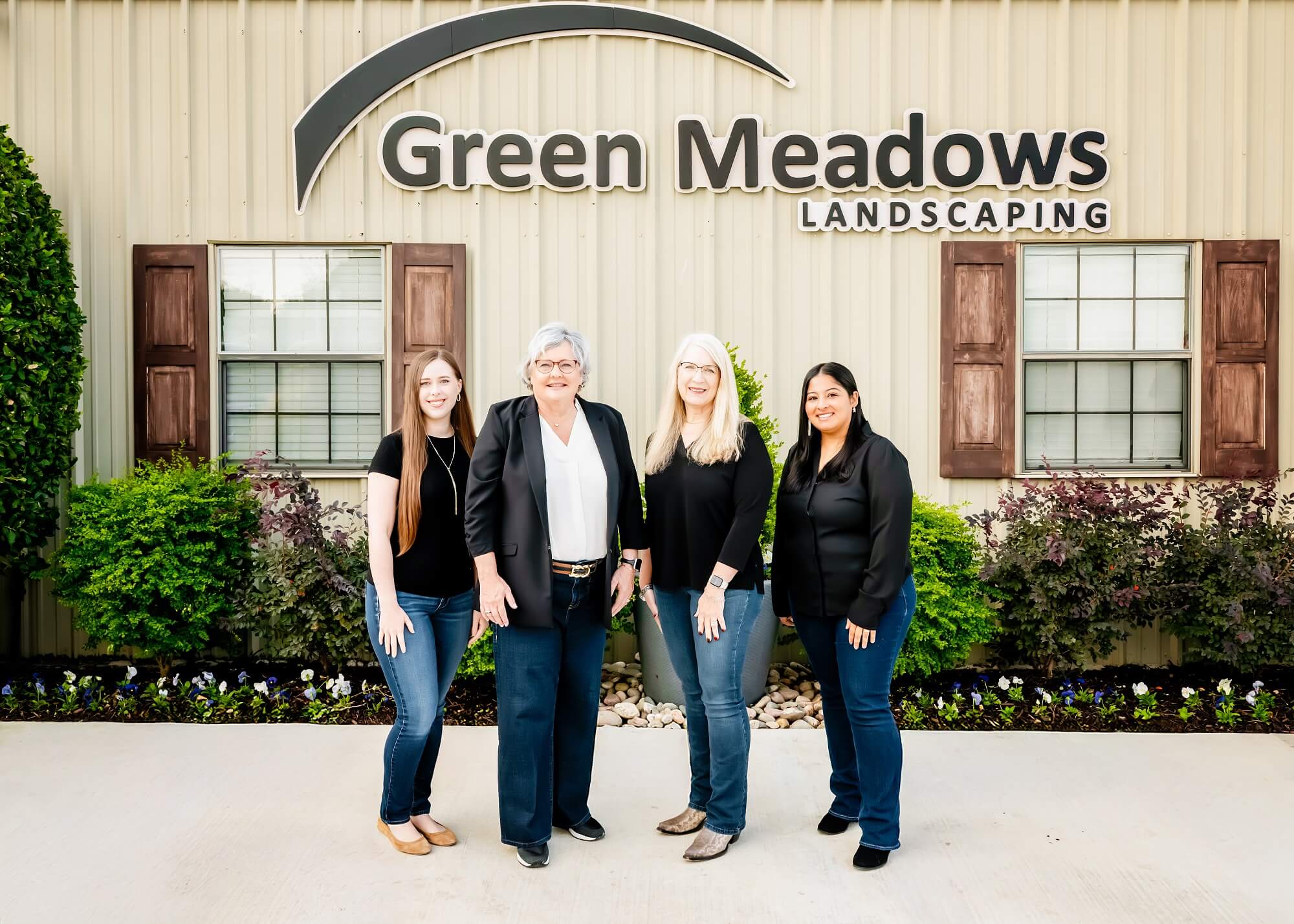 Four women stand smiling in front of a building with the sign Green Meadows Landscaping, flanked by windows with brown shutters and greenery, on a bright, sunny day.