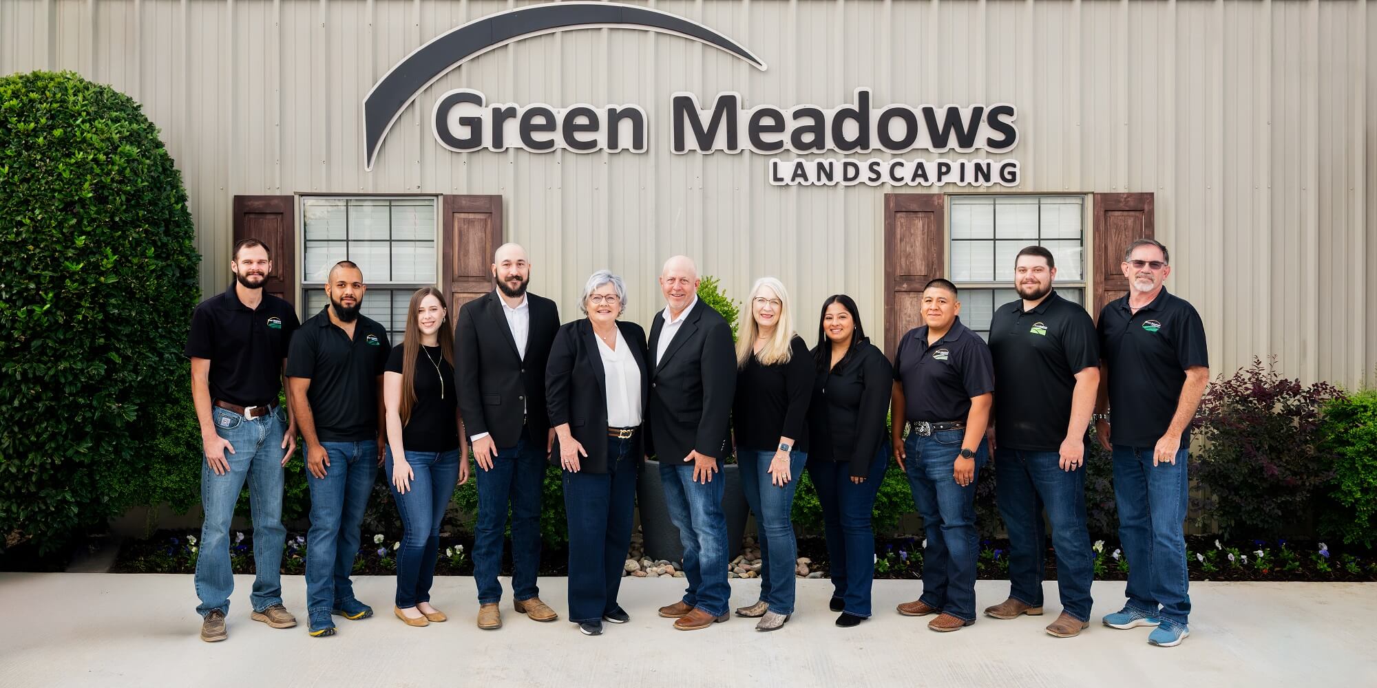Ten people stand smiling in front of a building with a Green Meadows Landscaping sign. They are dressed in business casual and pose together, suggesting they are staff or team members of the company. Shrubs decorate the background.