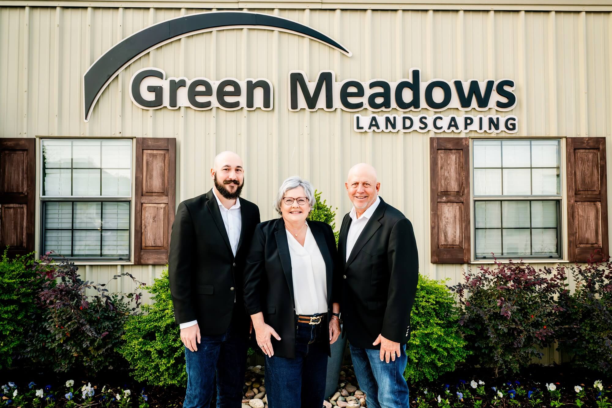 Three people stand smiling in front of a building with a sign that reads Green Meadows Landscaping. They are wearing business casual outfits and are surrounded by greenery and landscaping.
