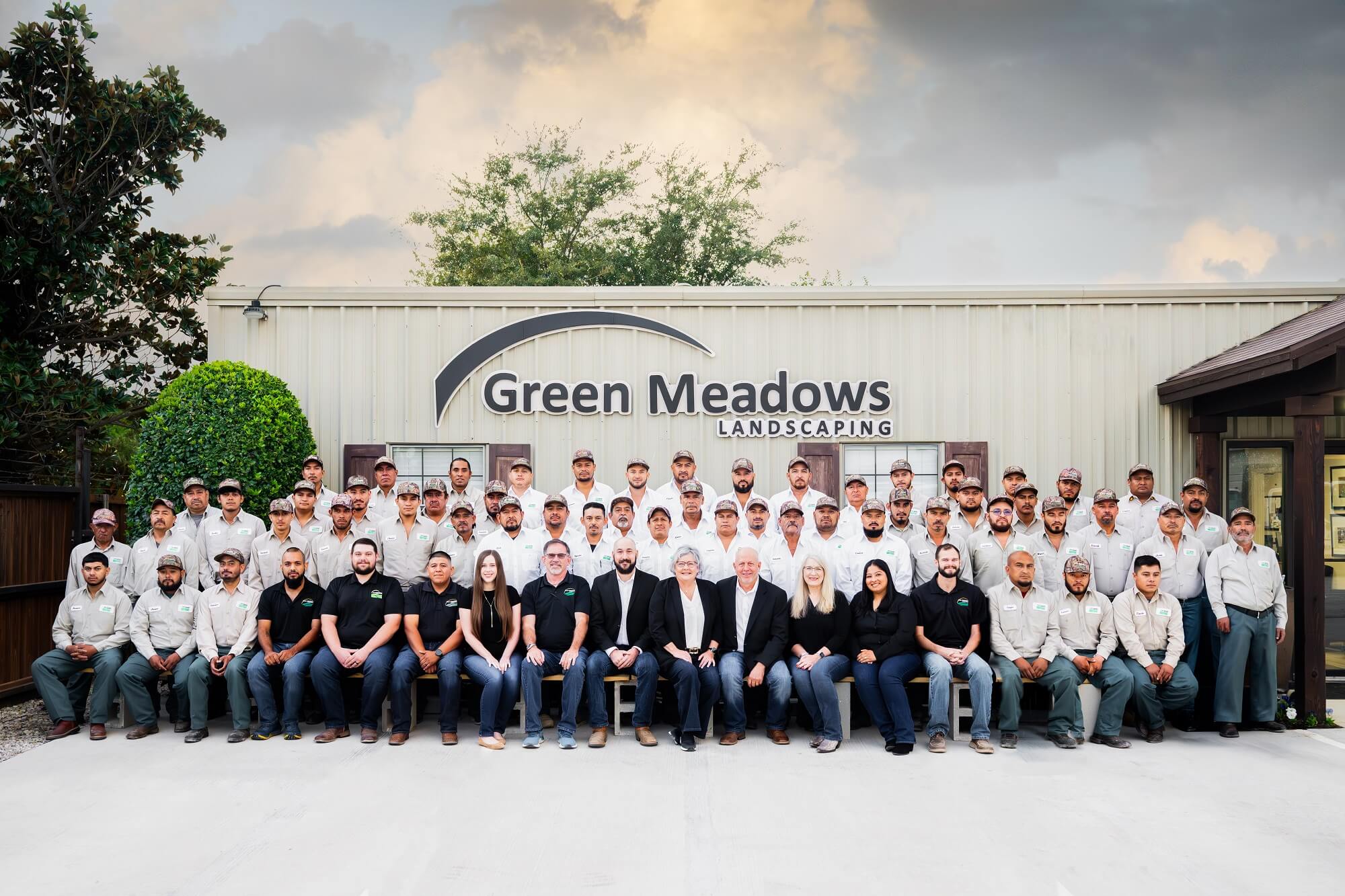A large group of people, mostly in uniforms, pose for a group photo in front of a building with a sign that reads Green Meadows Landscaping. Some are seated in front while others stand behind them.